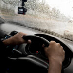 Hand of driving man use car steering wheel in side of car. Driving a leafy car on a rainy road. Car glass full of rain drops. Rural landscape with trees on the side of the road.